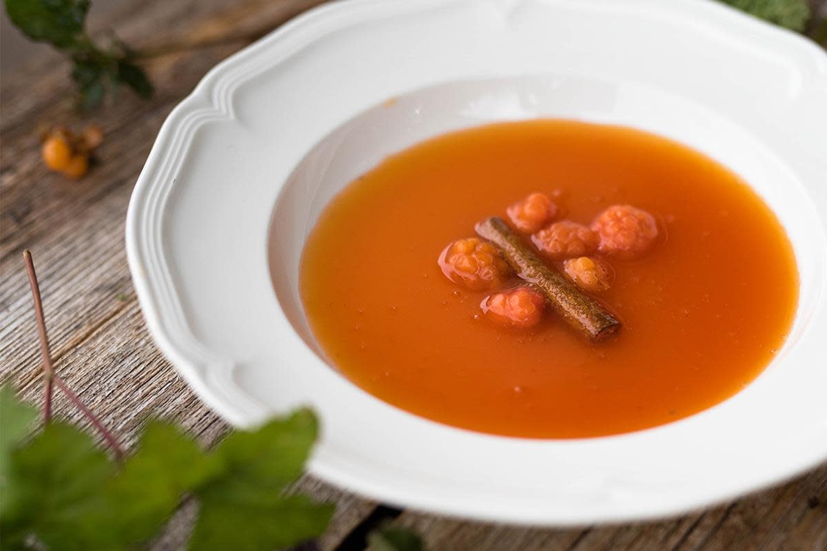 Krydret oransje multesuppe servert i hvit klassisk suppeskål, med hele multer og kanelstang. Står på rustikk trebord, pyntet med grønne blader.