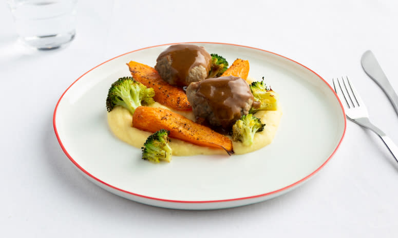 Kjøttkaker med bakt brokkoli og gulrøtter, potetmos og brun saus, servert på en hvit tallerken med rød kant.