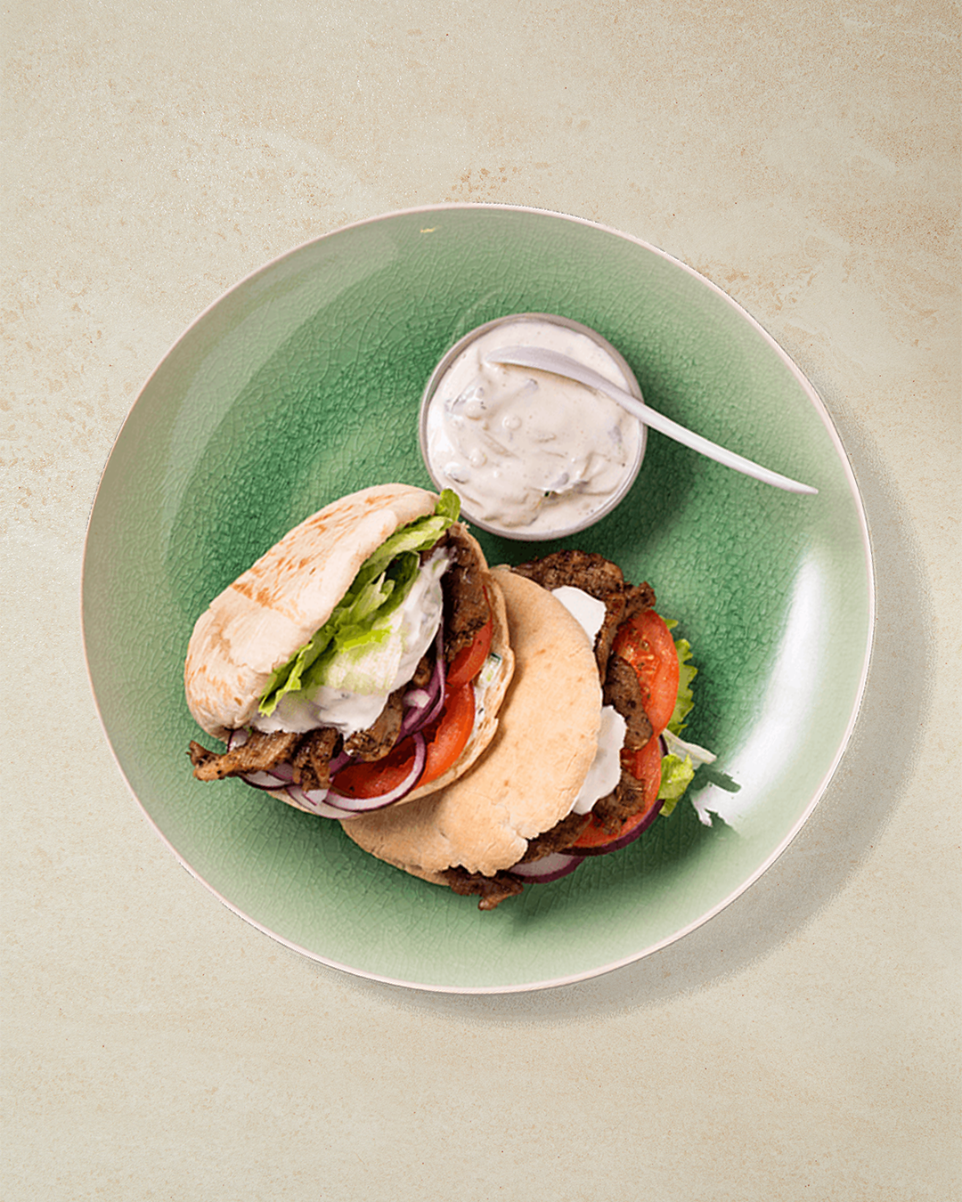 Gyros med svinekjøtt servert i pitabrød med salat, rødløk, tomater og tzatziki servert på grønn tallerken. Hjemmelaget tzatziki i liten skål ved siden av på tallerkenen.