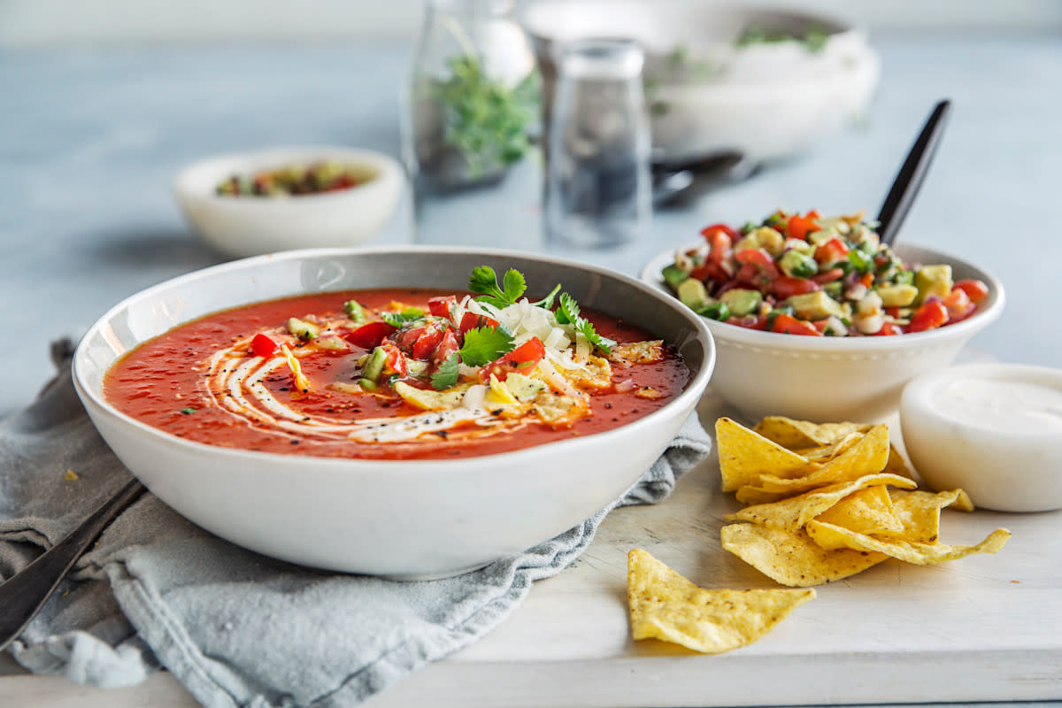 Fyldig tacosuppe med paprika, bønner og frisk avokadosalsa servert i lys skål, med avokado, paprika, ost og urter på toppen. Skål med topping i bakgrunnen, og tortillachips på siden.
