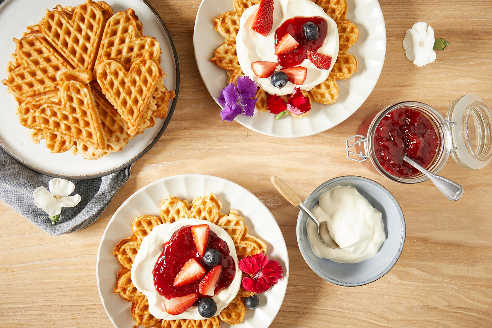 Glutenfrie vafler servert på hvite tallerkner på lyst trebord, med rømme, syltetøy og friske bær. 