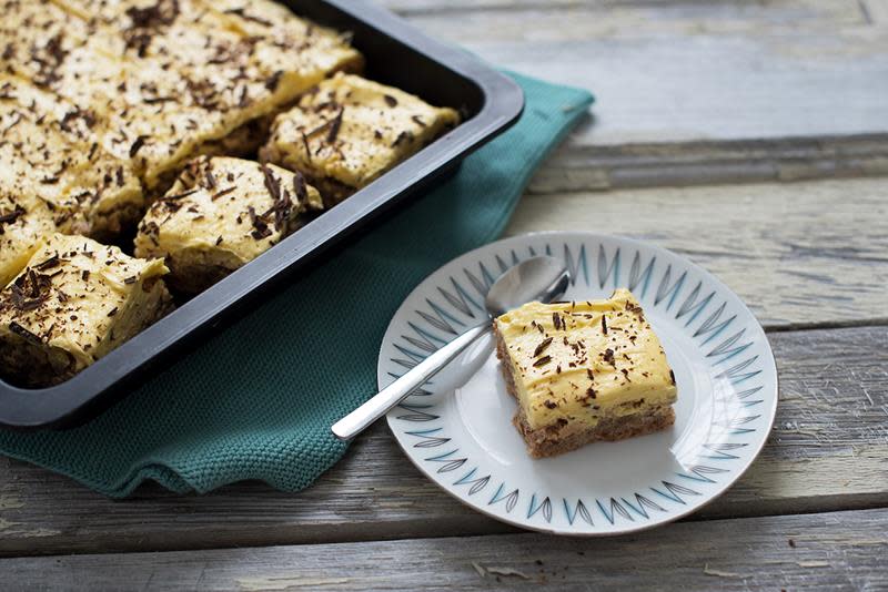 Suksessterte i langpanne, delt i biter, overstrødd med revet sjokolade og servert på rustikk trebord. En liten asjett med en bit suksessterte står ved siden av langpannen.