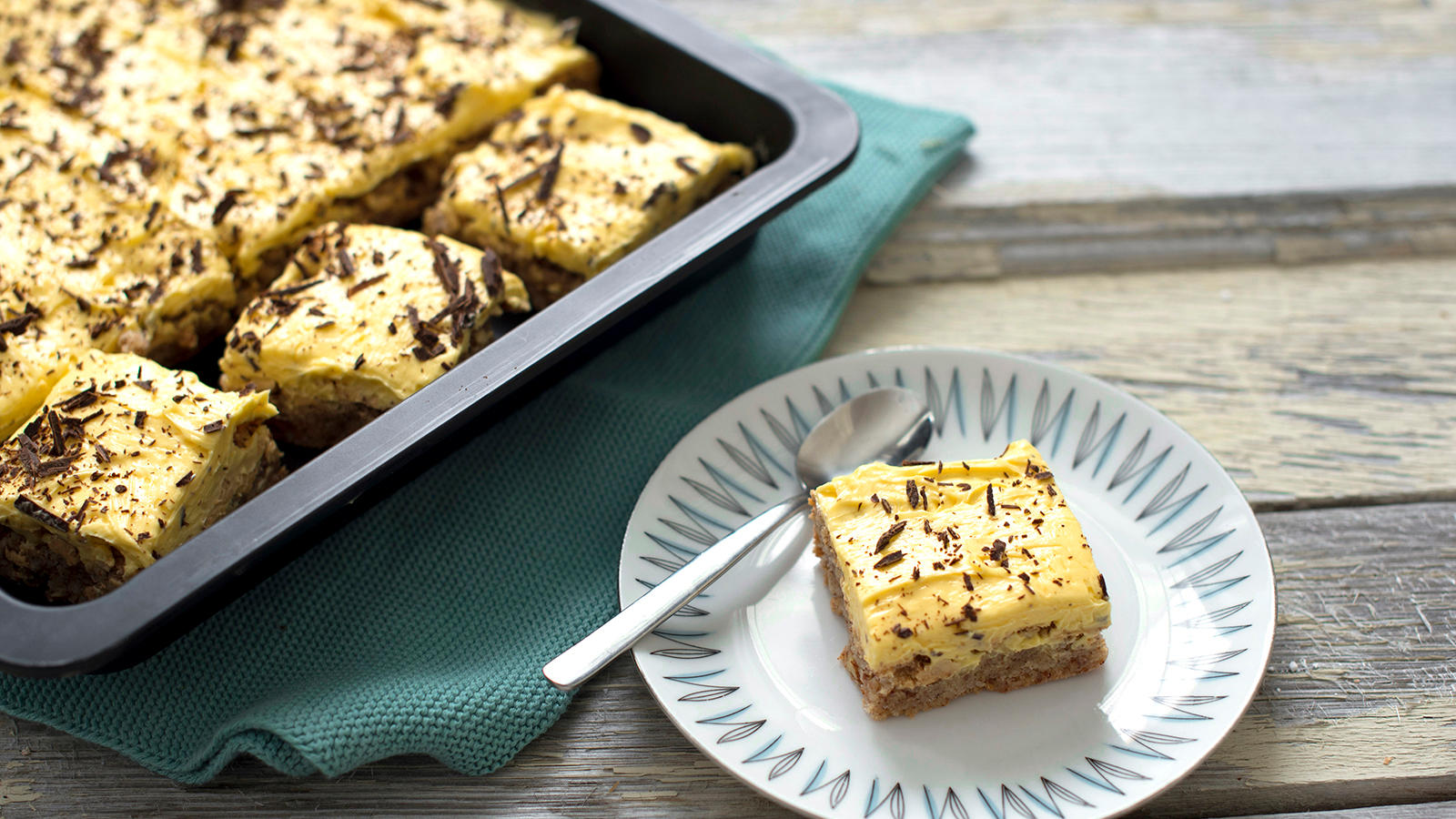 Suksessterte i langpanne, delt i biter, overstrødd med revet sjokolade og servert på rustikk trebord. En liten asjett med en bit suksessterte står ved siden av langpannen.