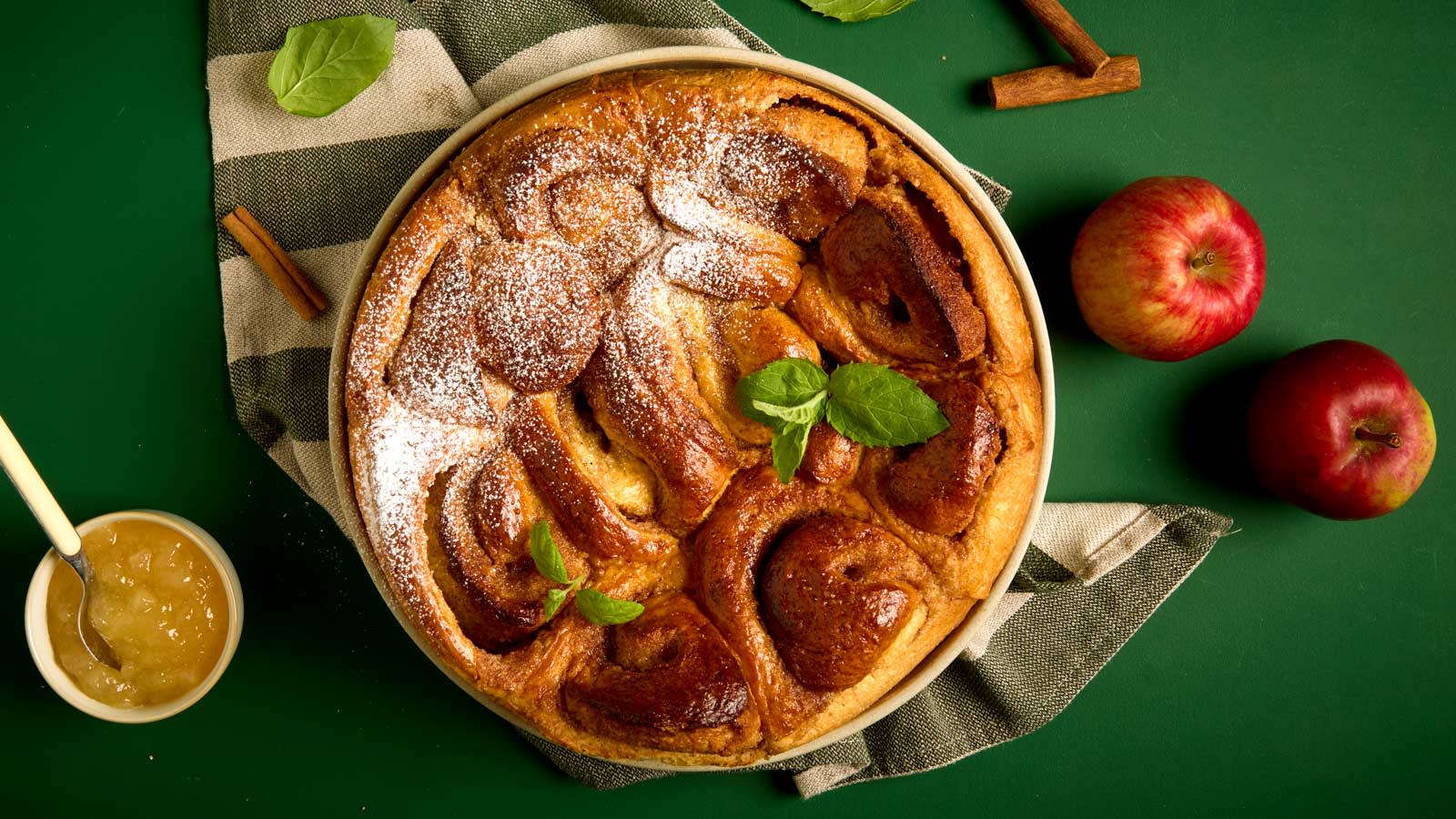 Kanelbollekake med eple og marsipan servet å grønt- og naturfarget kjøkkenhåndkle. Liten skål med eplesyltetøy ved siden av.