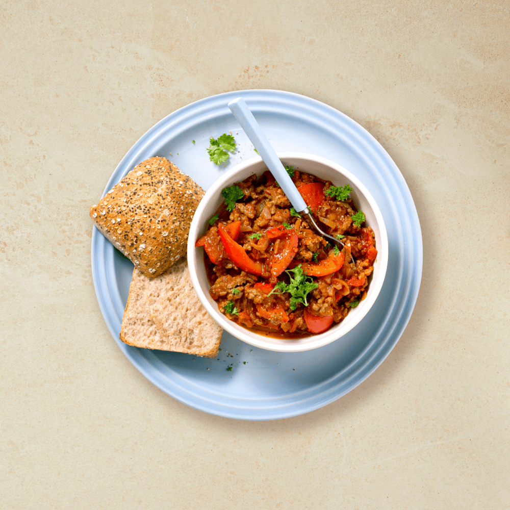 Gulasj med kjøttdeig i en liten hvit skål, det ligger et oppskjært rundstykke ved siden av, alt ligger på en lys blå tallerken