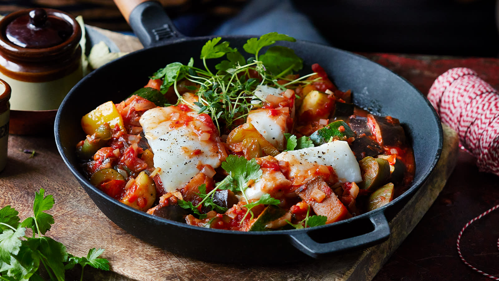 Ratatouille med torsk servert med friske urter, i mørk støpejernspanne på rustikk trefjøl.