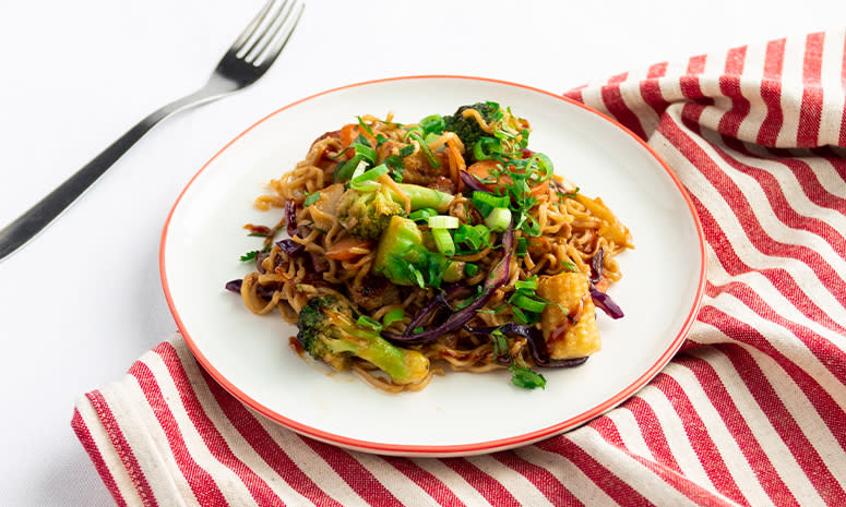 Wok med svinekjøtt servert på en hvit tallerken med røk kant, ligger på et hvitt og rødt stripete håndkle, det ligger en gaffel til venstre i bakgrunnen