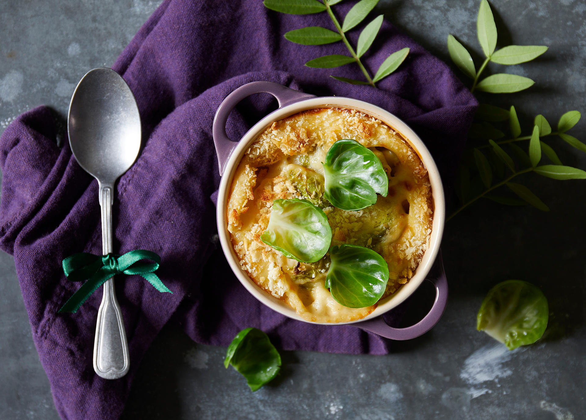 Rosenkål mac & cheese, makaronigrateng med rosenkål og sprøtt lokk, servert i lilla porsjonsform på en mørkelilla linserviett. Rosenkålblader på toppen, og en skje ved siden av.