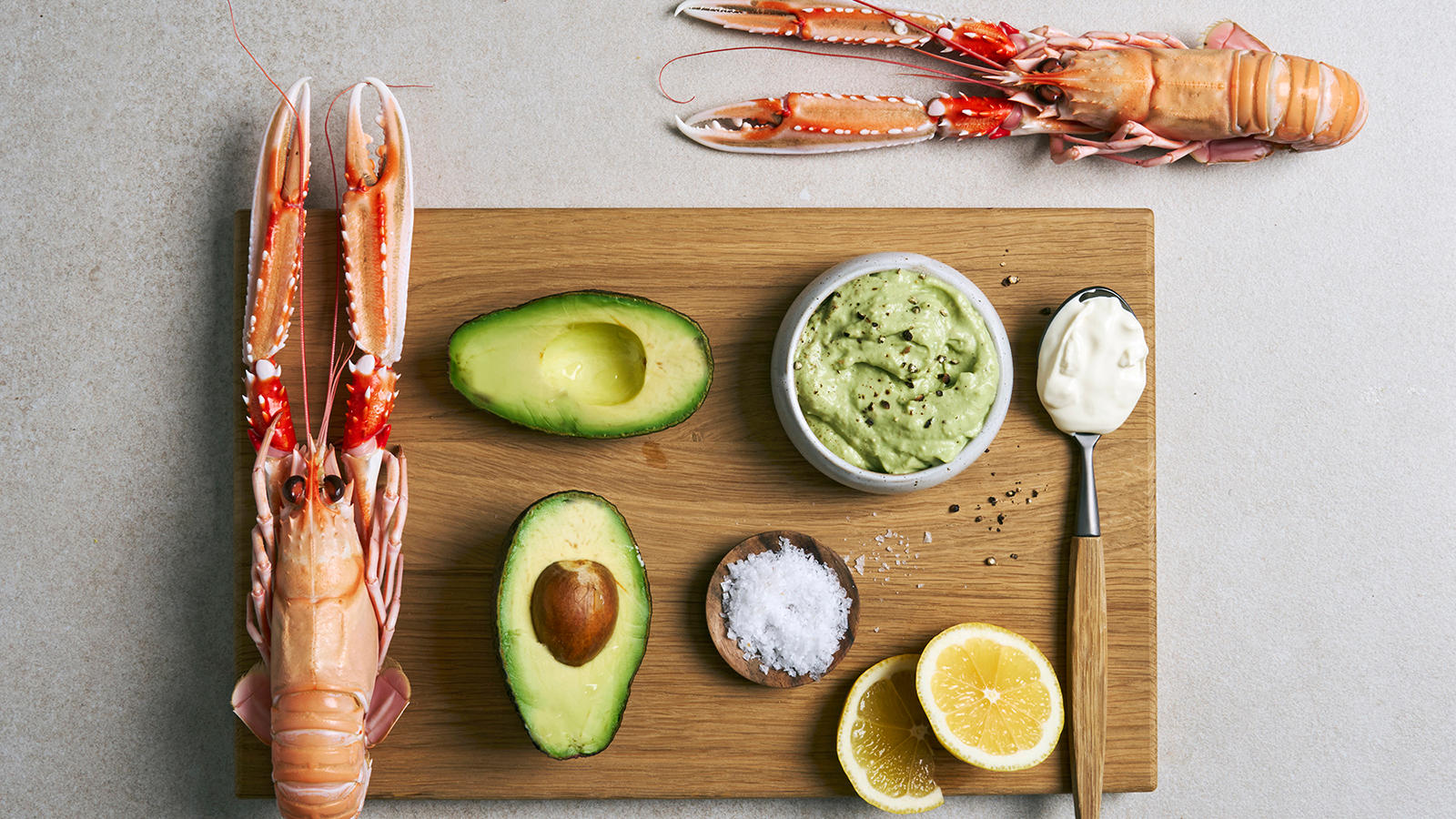 Avokadokrem servert i liten lys skål på trefjøl med frisk avokado, sitronskiver, crème fraîche og en hel sjøkreps.
