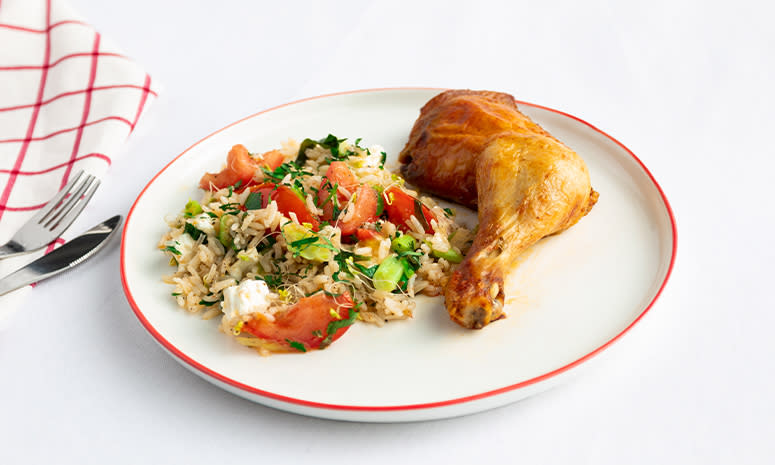 Kyllinglår med tomater, fetaost og ris, servert på en hvit tallerken med rød kant, det ligger bestikk på et rødt og hvitt håndkle til venstre for tallerkenen