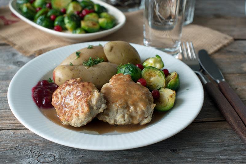 Hjemmelagede medisterkaker servert på hvit tallerken, med brun saus, poteter, rosenkål og tyttebærsyltetøy på rustikk trebord.