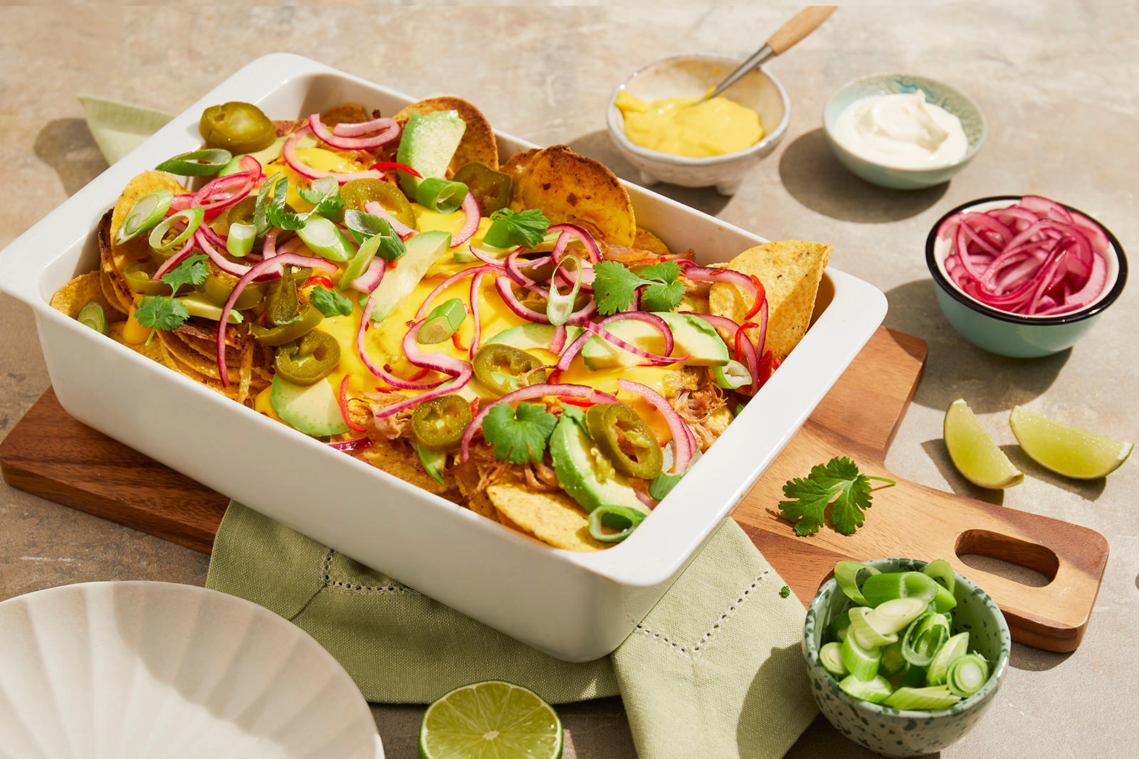 Nachos med pulled pork og kavli cheddar dipp servert i hvit ildfast form med jalapenos, avokado, rødløk og koriander på toppen. Små skåler med rømme, cheddar dipp, rødløk i skiver og vårløk i skiver rundt.