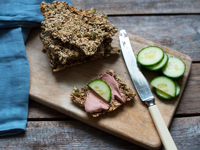Hjemmelagede frøknekkebrød servert på liten trefjøl, med blå linserviett halvveis over. Ett av knekkebrødene er påsmurt leverpostei og agurk, med ekstra agurkskiver på siden.