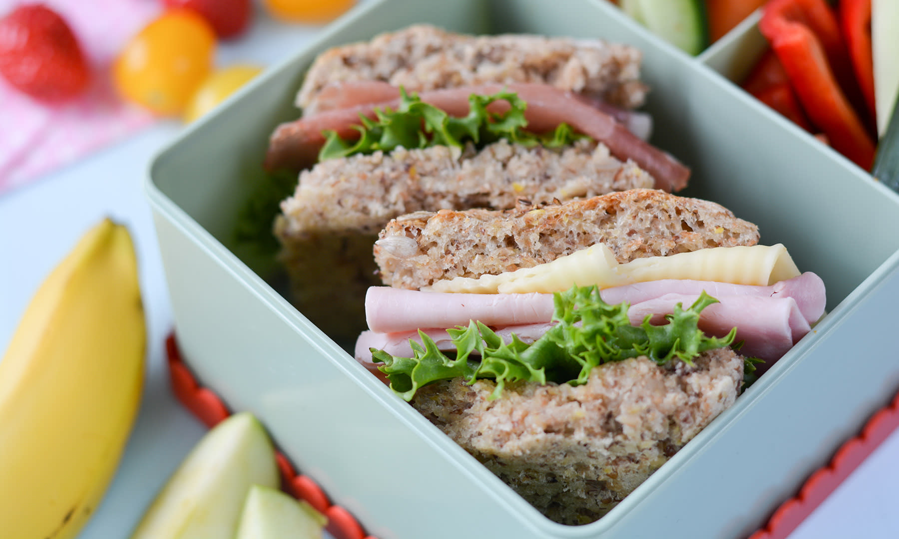 Grovt brytebrød i langpanne delt i to med ost, skinke og salat mellom, og servert i matpakke med banan, eplebåter og små tomater ved siden av.