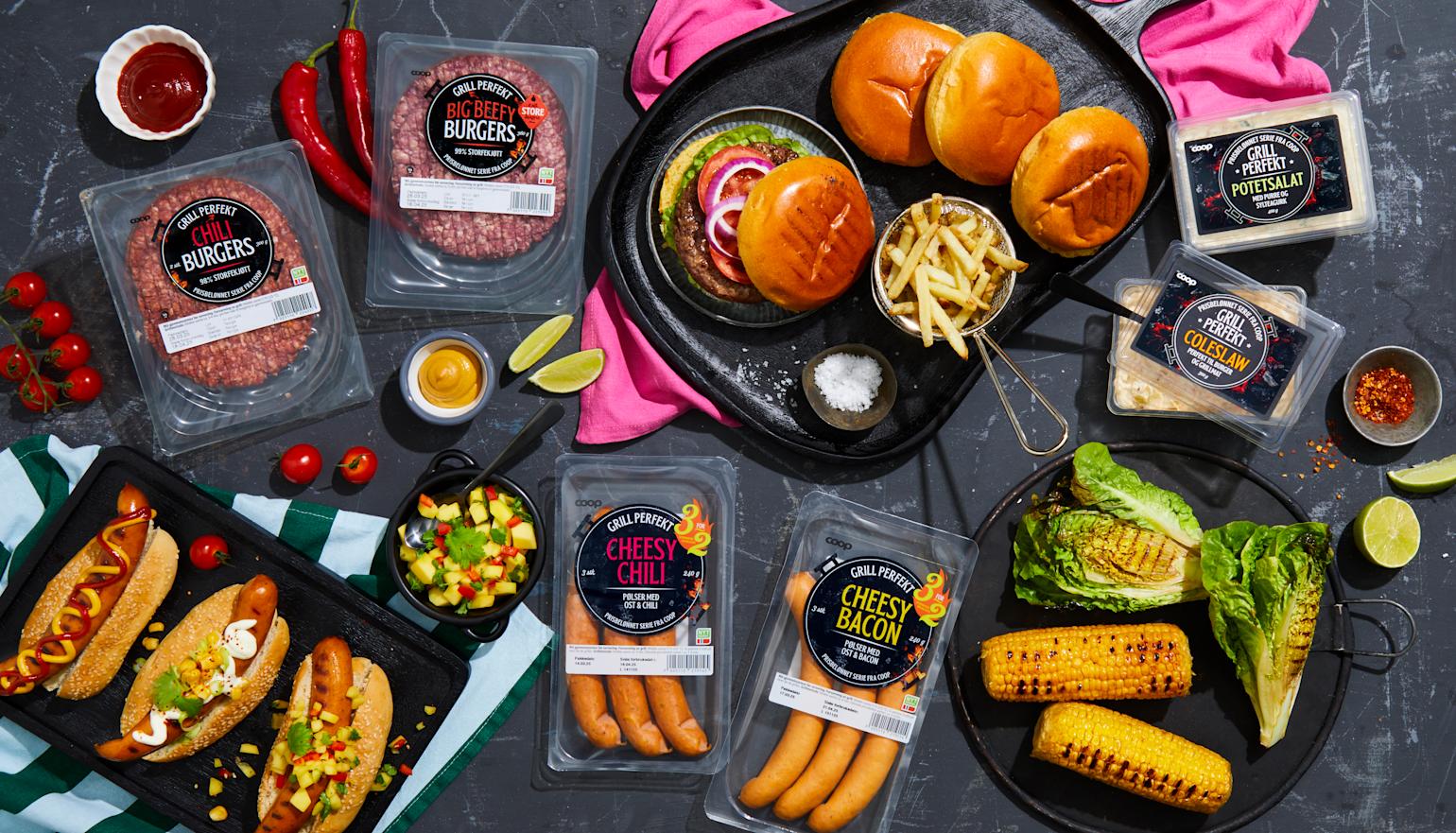 Grill Perfekt burgere og pølser i emballasje på mørk steinbakgrunn, samt tilberedt på svarte serveringsfat. Grillede maiskolber og grillet hjertesalat, og Grill Perfekt tilbehør ved siden av.