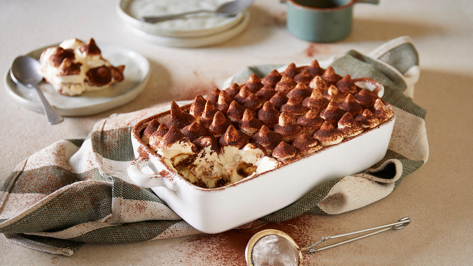Italiensk tiramisu servert i hvit ildfast form med et dryss mørk bakekakao. Formen står på et hvitt og grønt kjøkkenhåndkle. Asjetter med tiramisu i bakgrunnen.