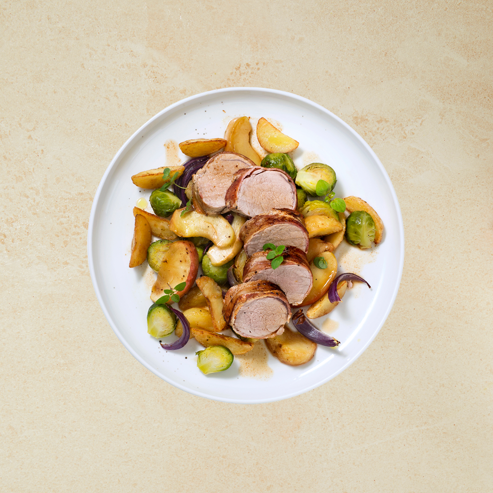 Baconsurret svinefilet med epler, grønnsaker og potetbåter servert på hvit tallerken med rødløk og et dryss ferske urter.