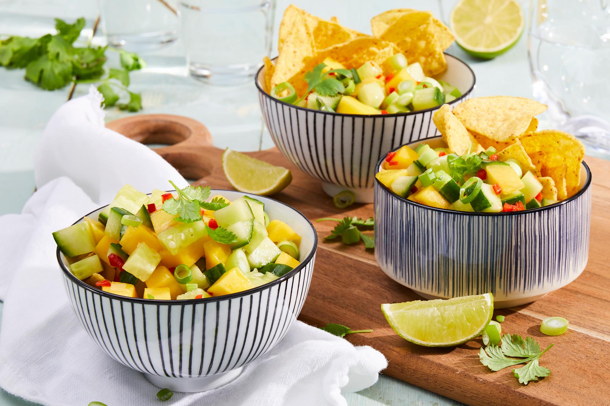 Mangosalsa med chili og agurk servert i tre forskjellige stripede skåler på trefjøl med frisk koriander og limebåter rundt. Vannglass i bakgrunnen.