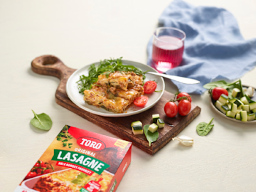 Lasagne på en tallerken som står oppå en fjøl med oppkuttet squash og hele tillater rundt seg med en Toro lasagne boks i forgrunnen