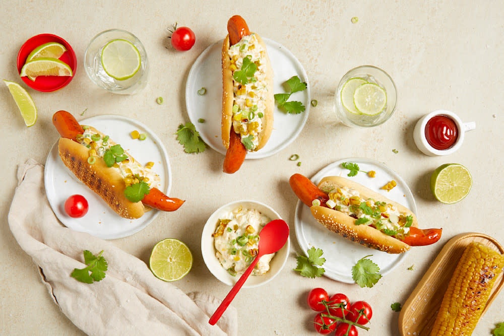 Spicy chicken pølse med meksikansk maissalat servert på tre små hvit asjetter på beige bakgrunn, med halve lime, frisk koriander og cherrytomater rundt. En grillet maiskolbe på siden.