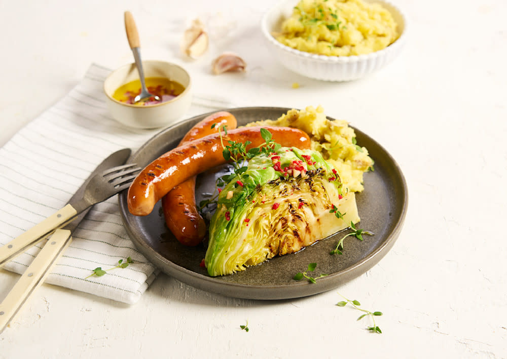 Cheesy bacon pølse med hjemmelaget potetmos og grillet nykål med hjemmelaget grillolje, servert på mørkegrå tallerken. Skåler med grillolje og potetmos i bakgrunnen.