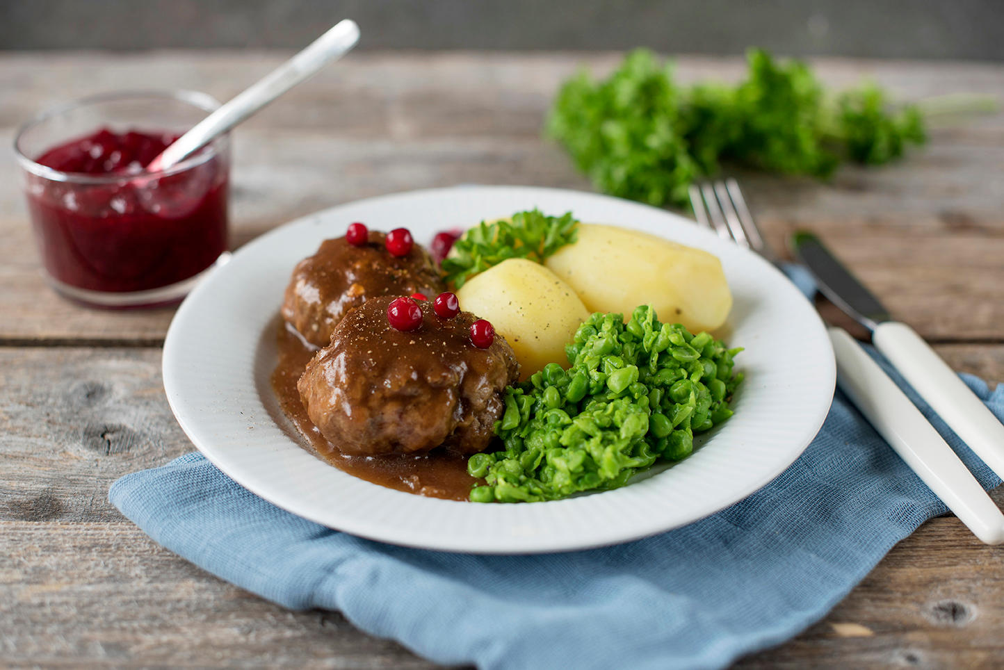 Kjøttkaker med brun saus, hjemmelaget ertestuing og kokte poteter. Tyttebær på toppen. Servert på en hvit tallerken, på rustikk trebord.