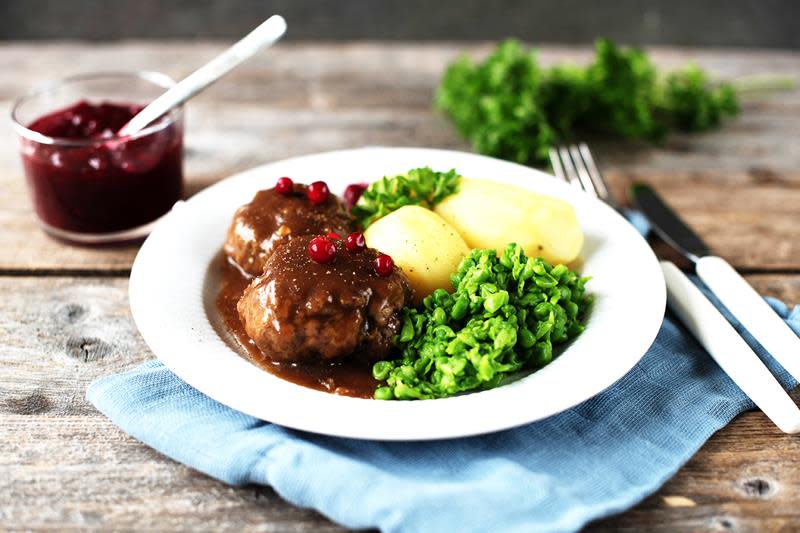 Kjøttkaker med brun saus, hjemmelaget ertestuing og kokte poteter. Tyttebær på toppen. Servert på en hvit tallerken, på rustikk trebakgrunn.