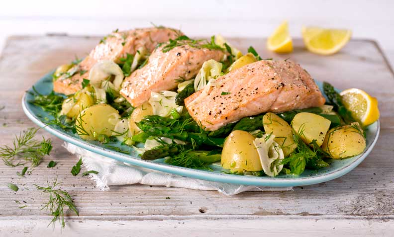 Ovnsbakt laks med potet-og urtesalat med fennikel og asparges, på turkist fat. Lys trebakgrunn, ferske sitronbåter og friske urter på siden.