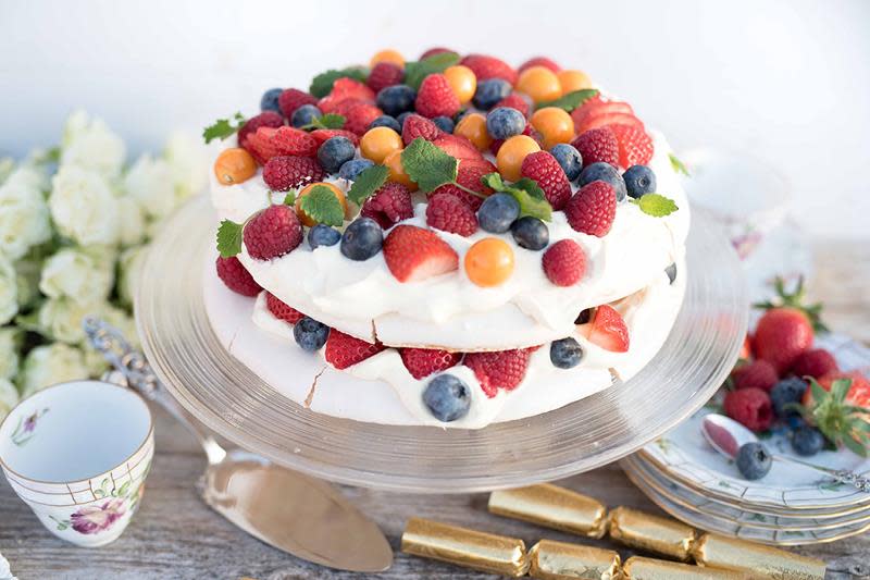 Pavlova med hjemmelaget vaniljekrem og friske blåbær, bringebær, jordbær og physalis, servert i to lag på stettfat. Fatet står på hyggelig dekket kaffebord med blomster og festpynt rundt.