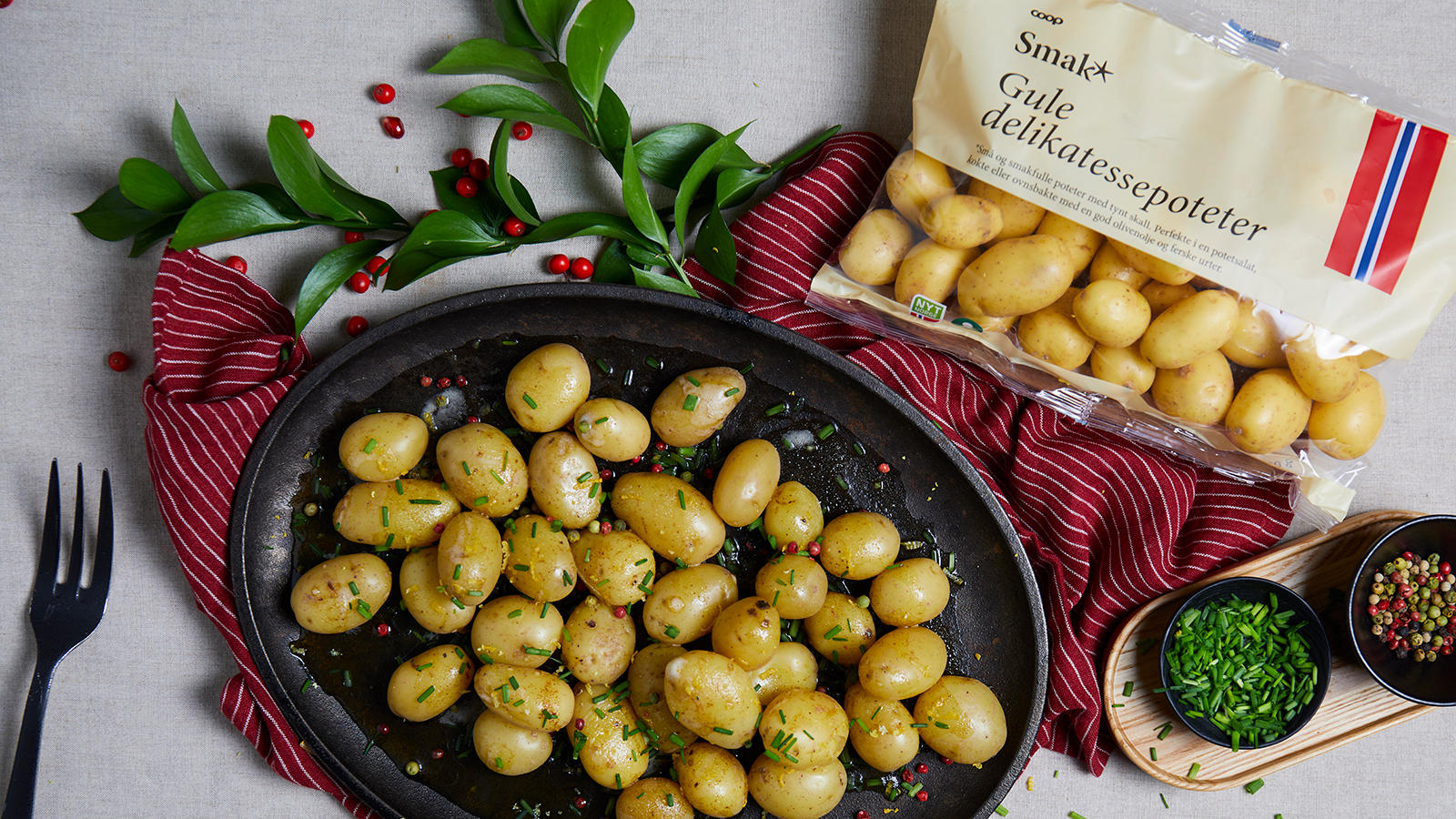 Delikatessepoteter med brunet smør og sitron servert på ovalt svart fat med gressløk og pepperkorn over. Rødt kjøkkenhåndkle med hvite striper under, og små skåler med gressløk og pepperkorn på siden.