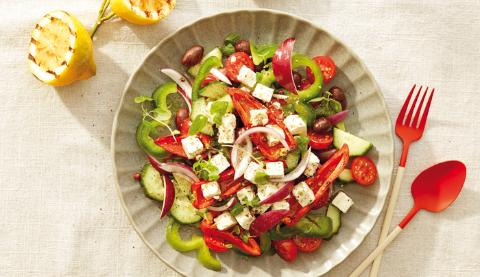 Gresk salat i en skål med rødt bestikk på høyre side av tallerkenen, servert med sitron.