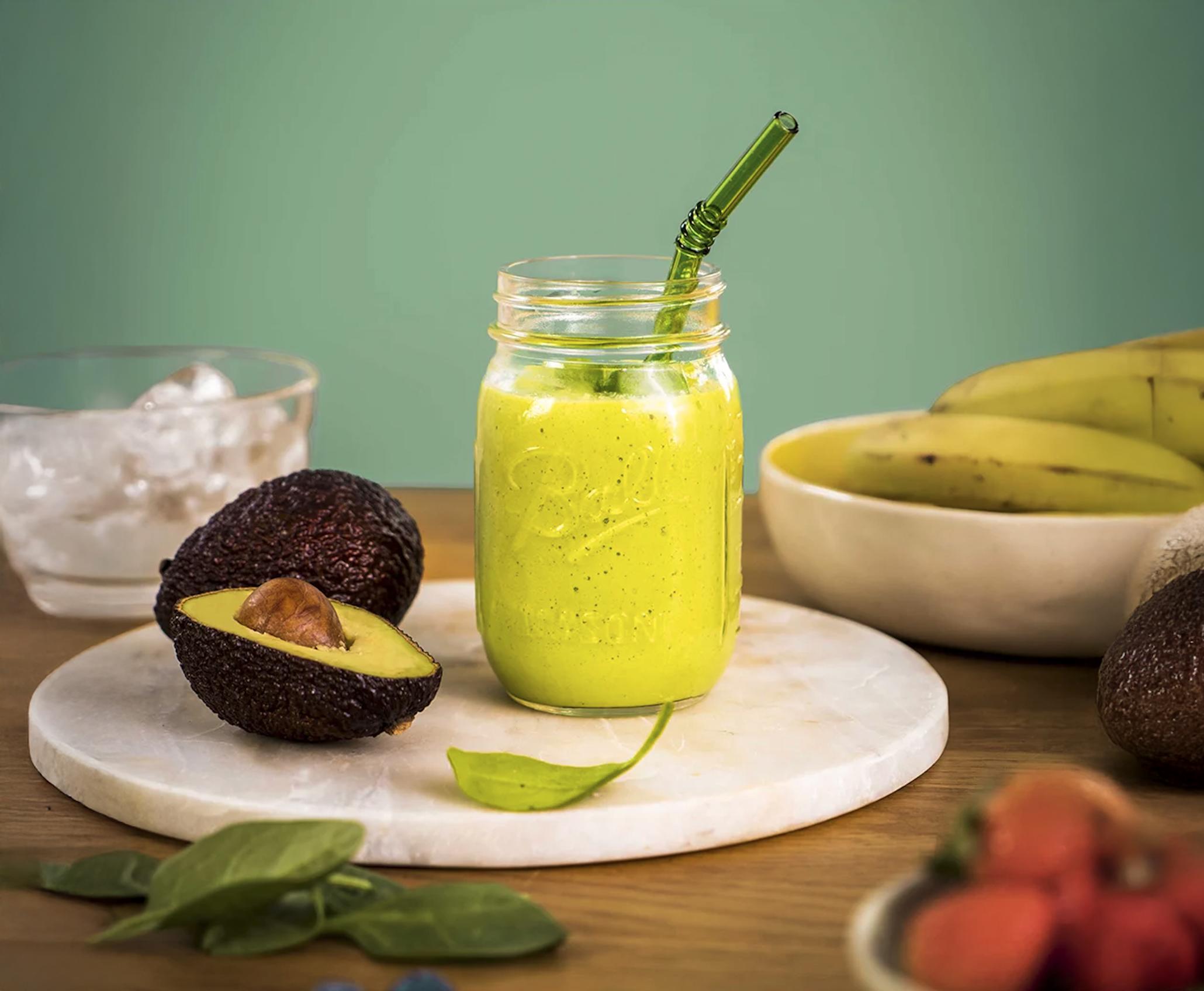 Sommergrønn smoothie med avokado, banan og spinat, i glass på hvit marmorfjøl med avokado, banan og spinat rundt.