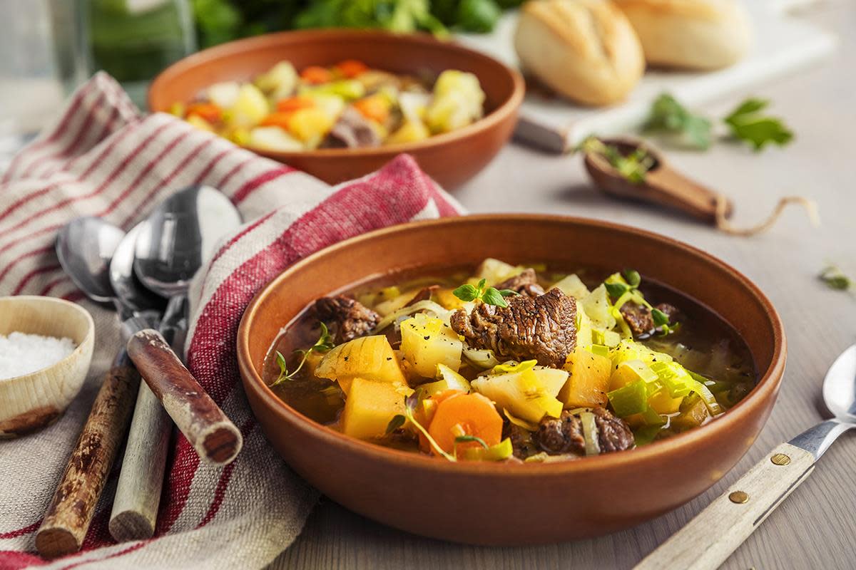 Kjøttsuppe med fargerike rotgrønnsaker, toppet med fersk timian, servert i mørk brun suppeskål. Beige og rødstripet linduk, ferske urter og rustikke skjeer ved siden av.