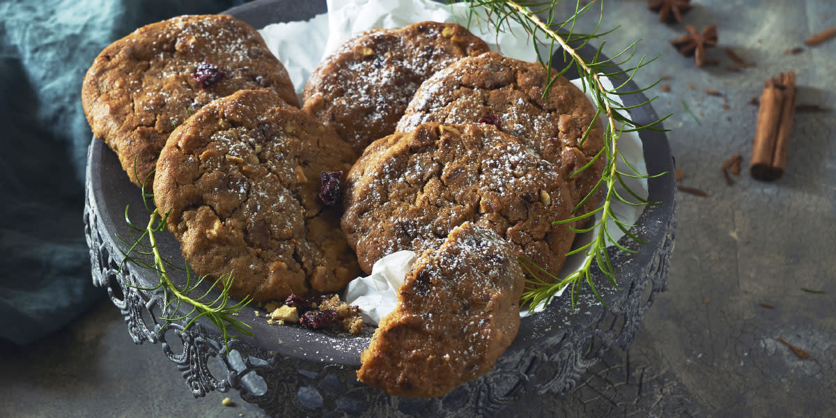 Julecookies med sjokolade, tranebær og peanøtter med melisdryss servert på hvitt bakepapir på mørkegrått kakefat i metall. Grønne kvister og hele tørkede krydder rundt.