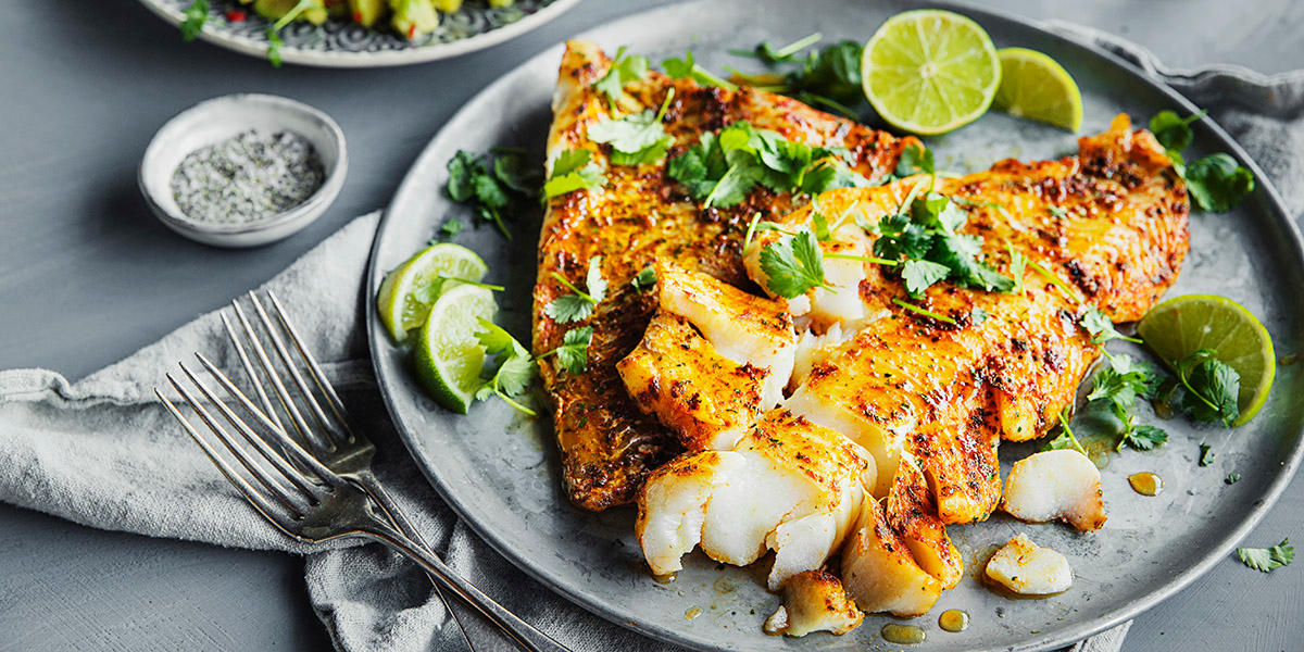 Spicy ovnsbakt skrei med krydret marinade servert på grå tallerken med koriander og limebåter. Tallerken med mango- og avokadosalat i bakgrunnen.