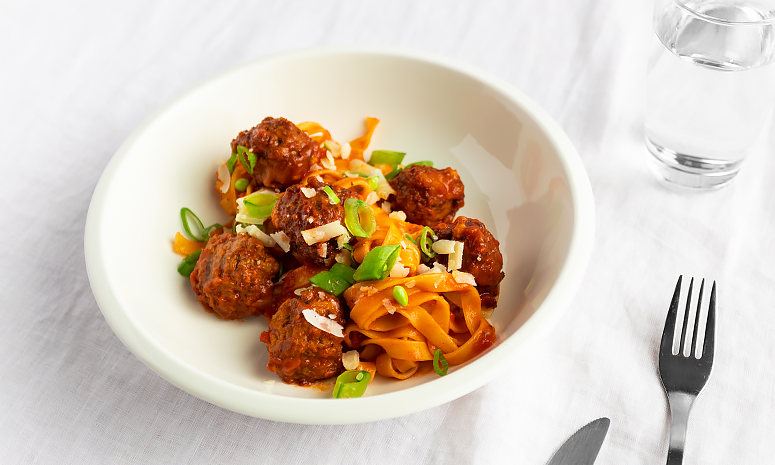 Kjøttboller og pasta i tomatsaus i en dyp hvit skål, bestikk ligger på høyre siden av skålen og det står et glass vann i bakgrunnen, alt er på en hvit duk