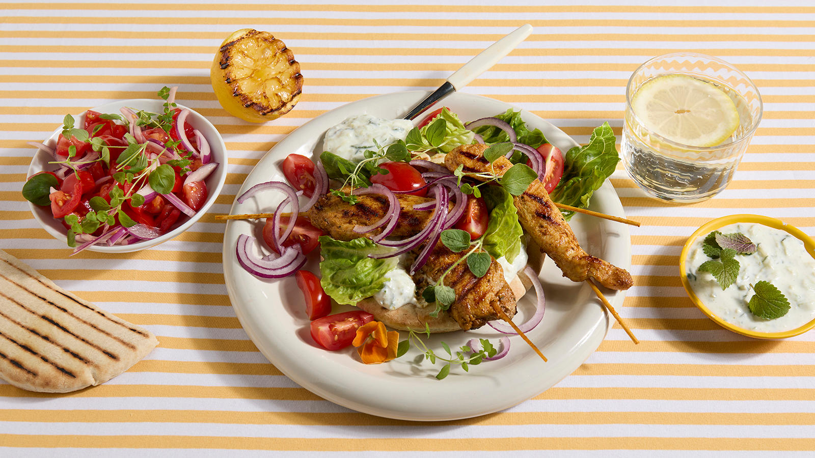 Kyllingspyd med jukse-tzatziki servert på lys tallerken med pitabrød, rødløk, cherrytomater og salat. Skåler med tilbehør rundt.