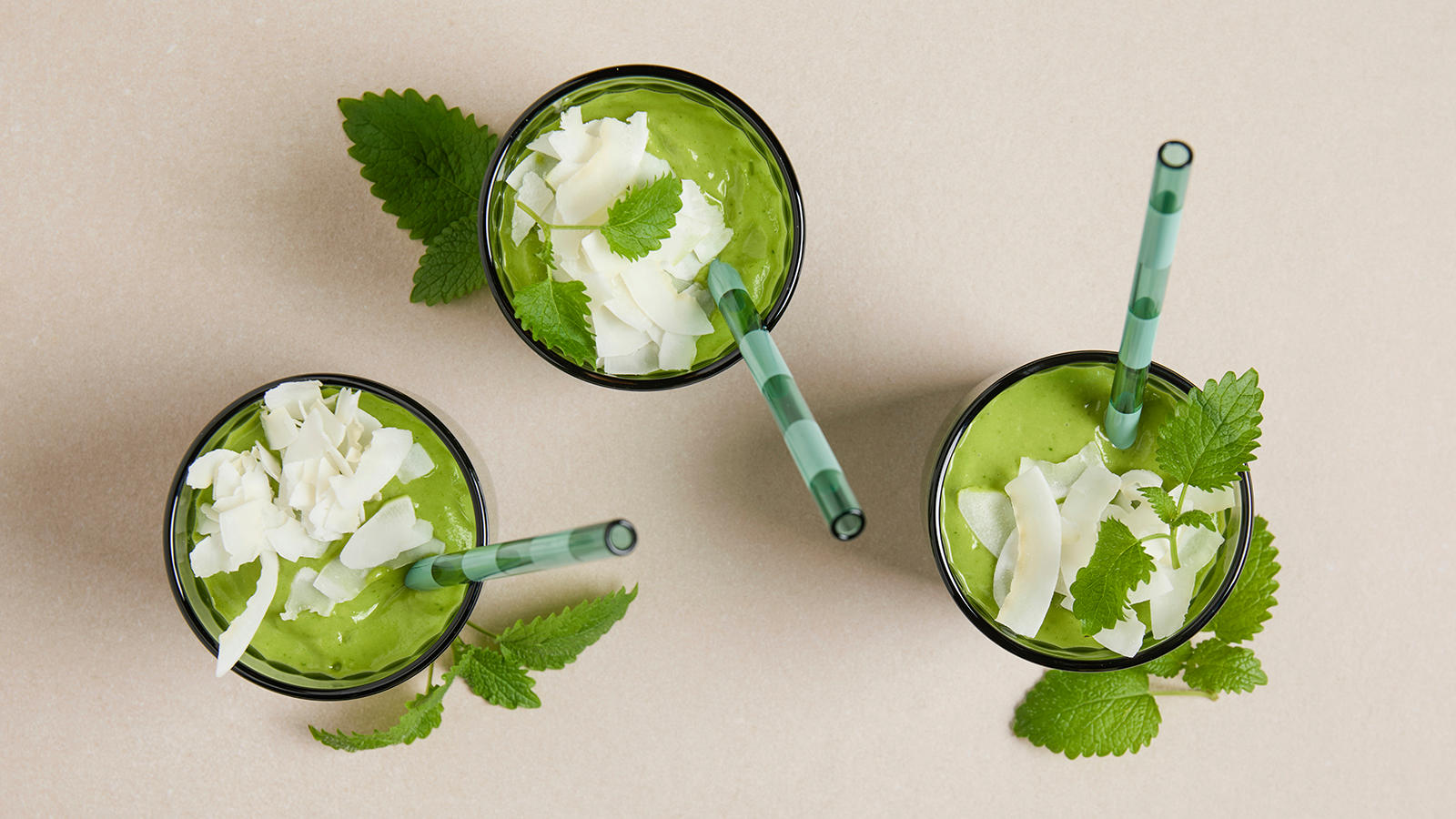 Grønn smoothie med avokado og tropisk smak servert i tre grønne glass med stripete sugerør, kokosflak og sitronmelisse.