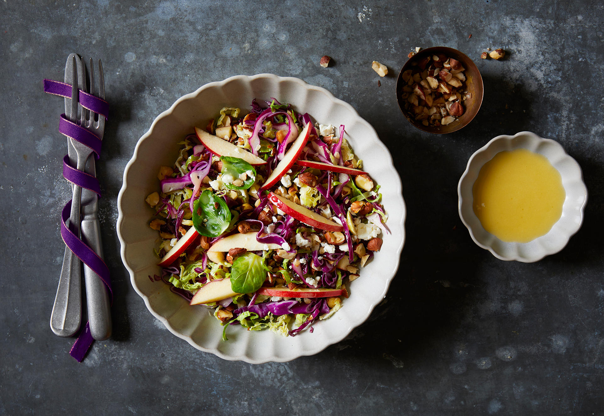Rosenkålsalat med sennepsvinaigrette, toppet med rødt eple i skiver, fetaost og rødkål i strimler, servert i hvit skål. Skålen står på mørkegrå bakgrunn, med to små skåler vinaigrette og grovhakkede nøtter ved siden av.