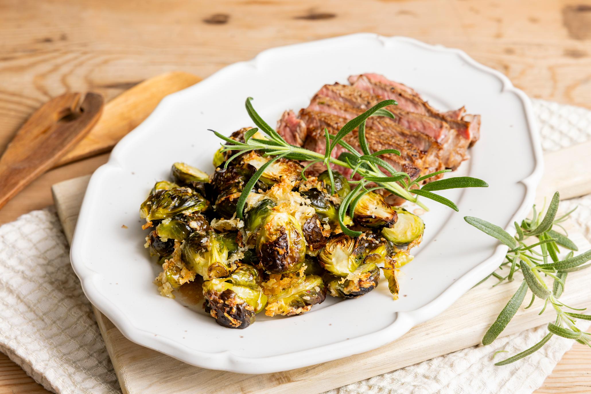 Crispy rosenkål med parmesan servert på hvitt ovalt fat sammen med et stykke stekt kjøtt, med en kvist frisk rosmarin. Fatet står på en lys trefjøl på kjøkkenhåndkle, på trebord.