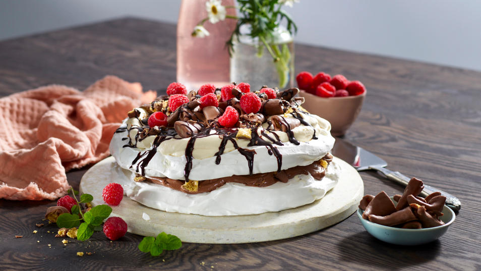 Pavlova Smashkake i to lag på hvitt kakefat, pyntet med sjokoladekrem, sjokoladesaus, pisket krem, Smash og friske bringebær. Patentflaske med lyserosa saft, liten skål med friske bringebær, og krukke med markblomster i bakgrunnen.