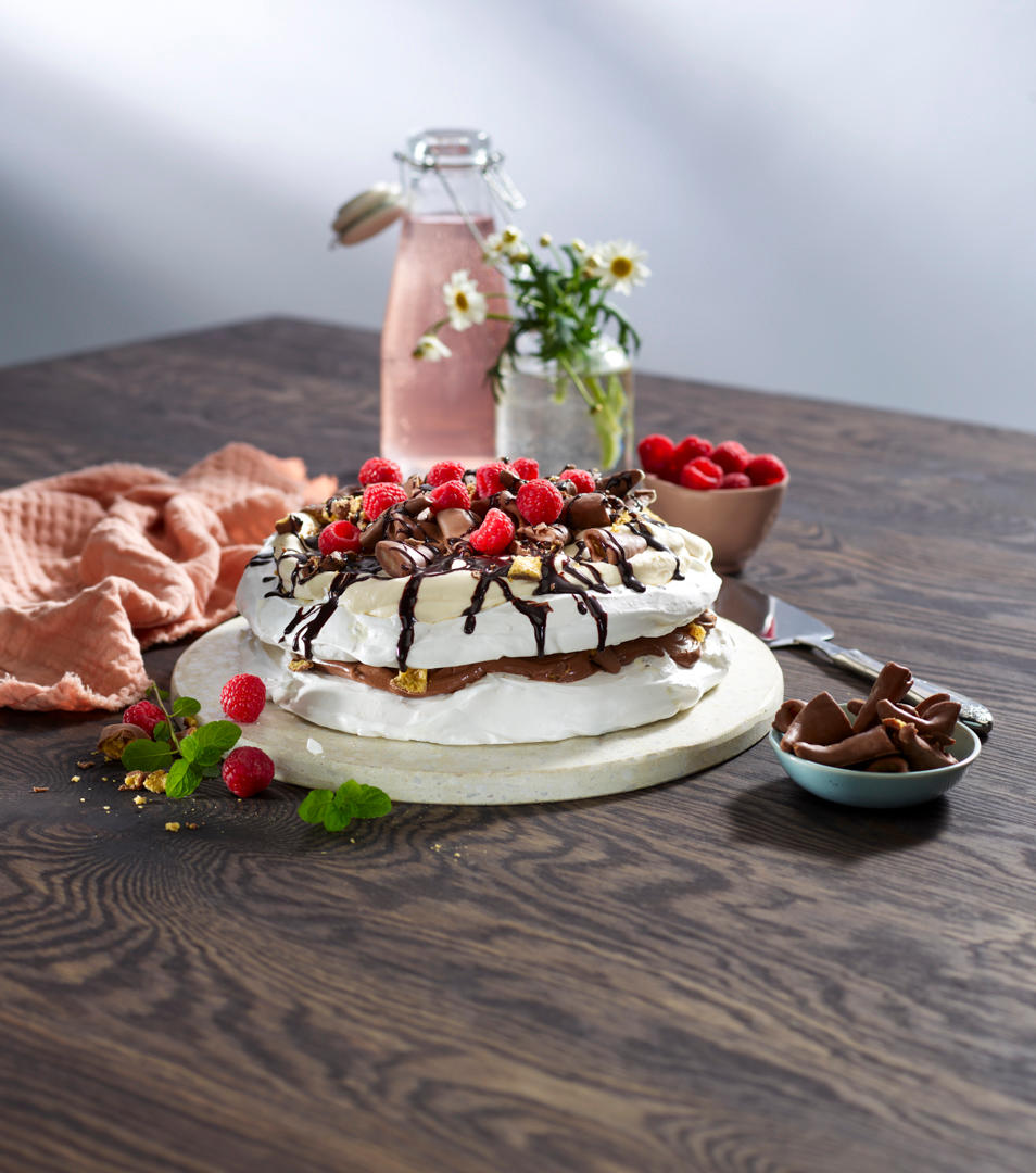 Pavlova Smashkake i to lag på hvitt kakefat, pyntet med sjokoladekrem, sjokoladesaus, pisket krem, Smash og friske bringebær. Patentflaske med lyserosa saft, liten skål med friske bringebær, og krukke med markblomster i bakgrunnen.