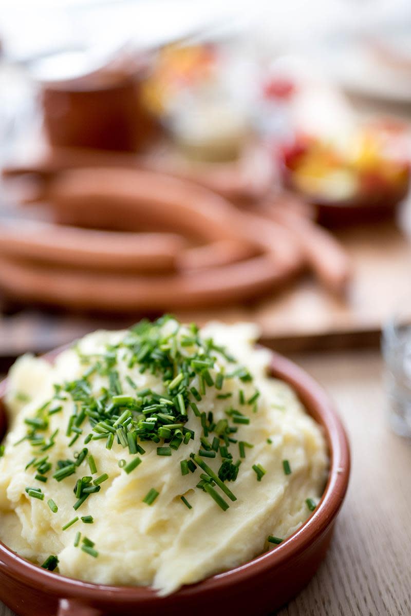 Enkel hjemmelaget potetmos, servert i mørk keramikkform, med et dryss gressløk. Kokte wienerpølser i bakgrunnen.