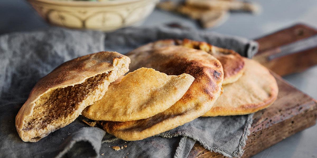 Grove pitabrød servert på mørk trefjøl med grå linserviett på. Bakebolle med mønster i bakgrunnen.