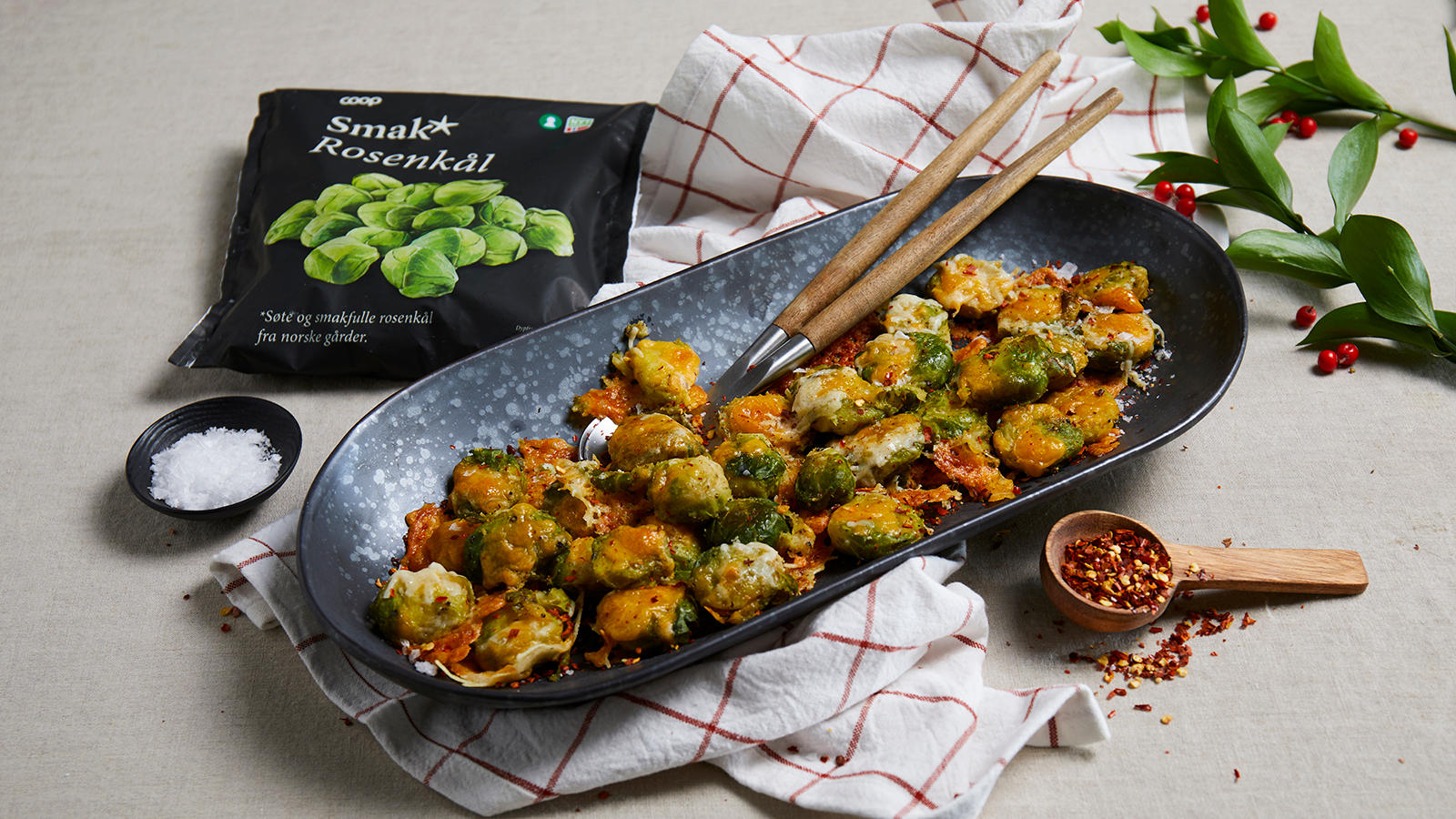 Smashed rosenkål med cheddar og parmesan drysset med chiliflakes og servert i avlang svart skål på hvitt og rødt kjøkkenhåndkle. Liten skål med flaksalt,og spiseskje i tre med chiliflakes ved siden av.