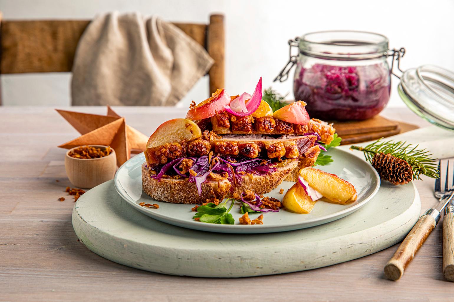 En skive røstet landbrød, med rødkål, skiver av gylden ribbe og smørstekte epler, toppet med syltet rødløk. Servert på lys grønn tallerken på lyst rustikk trebord. Julepynt, granbar og krukke med syltet rødkål i bakgrunnen.