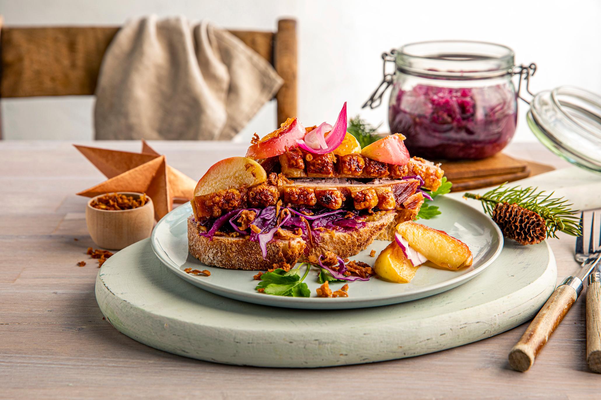 En skive røstet landbrød, med rødkål, skiver av gylden ribbe og smørstekte epler, toppet med syltet rødløk. Servert på lys grønn tallerken på lyst rustikk trebord. Julepynt, granbar og krukke med syltet rødkål i bakgrunnen.