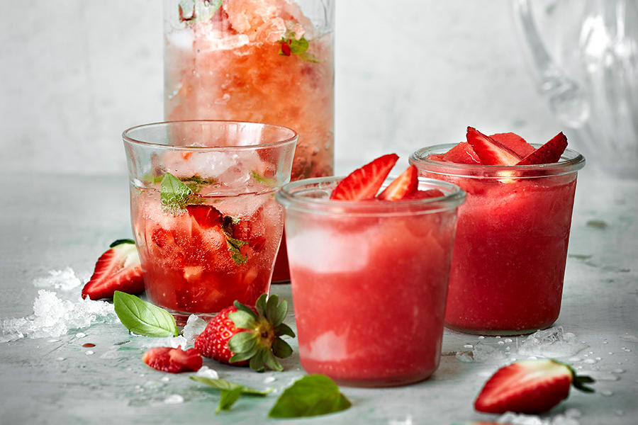 Jordbærmocktails og jordbærslush i glass på grått bord med knust is, friske jordbær og mynteblader rundt. Mugge med jordbærdrikk og isbiter i bakgrunnen.