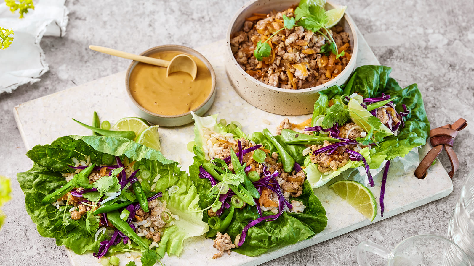 Hjertesalatwraps med peanøttdressing servert med grønnsaker og svinekjøttdeig på lys marmorfjøl. Liten skål med kjøttfyll, og liten skål med peanøttdressing ved siden av.