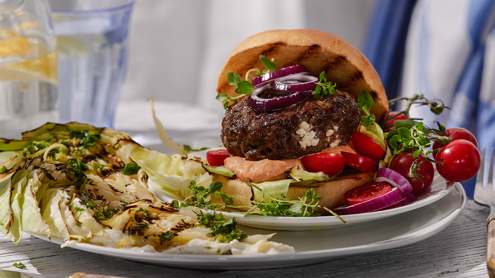 Gresk burger med grillet nykål og sitronyoghurt servert på hvit asjett med rødløk og piccolotomater.